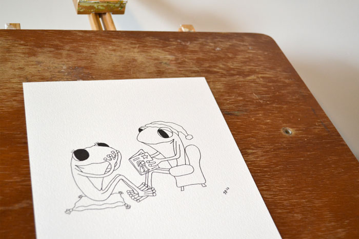 A photograph of an 'in progress' watercolour by Ruth Burton Artist for the Frog's Bucket List 'Christmas Fun' book. The image shows a pen outline on watercolour paper of Frog reading a Christmas story to Frog.