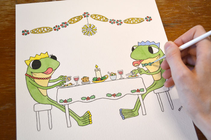 A photo of an 'in progress' watercolour, the image shows the watercolour paint being applied to an image of Frog and Frog eating Christmas dinner, by Ruth Burton Artist.