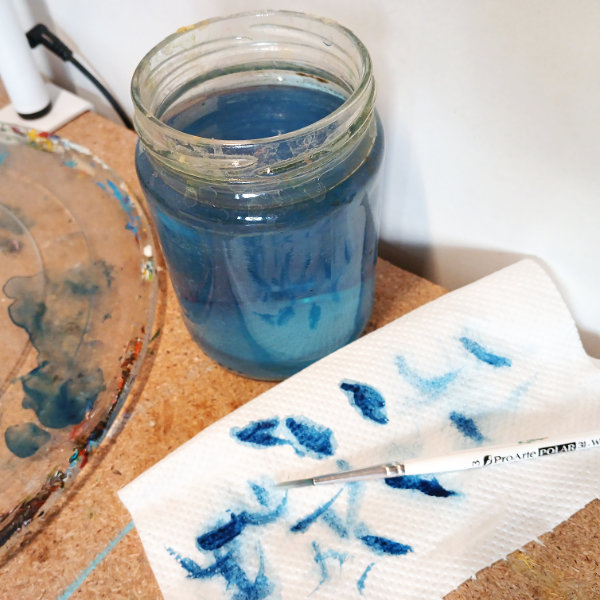 A photograph showing art materials at the end of the watercolour challenge. There is a jar of blue water, a paintbrush, a mixing palette and piece of tissue paper with blue paint on them.