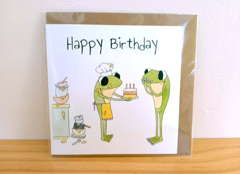 A frog birthday card by Ruth Burton. The illustration for the card is 'Frog's Birthday Cake' ('Bake a birthday cake'). The card is displayed with a kraft envelope in a cellophane bag.