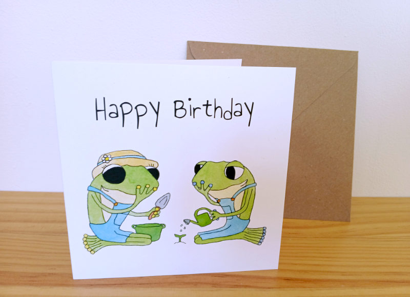 A frog birthday card by Ruth Burton. The illustration for the card is 'Plant a garden' and shows Frog and Frog planting a seedling. The card is displayed with a kraft envelope.