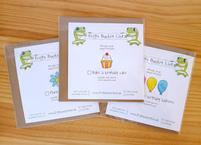 A photo of the reverse side of the three Frog's Bucket List greetings card samples by Ruth Burton Artist. The greetings cards show the tick box and 'bucket list' caption with other small illustrated elements (a flower, a cupcake, balloons).