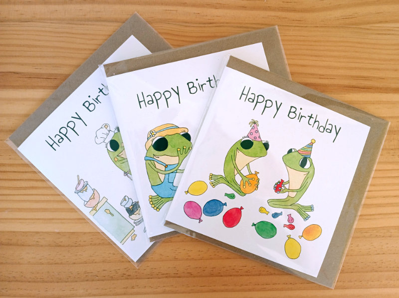 A photo of three Frog's Bucket List greetings card samples by Ruth Burton Artist. The greetings cards are arranged in a fan. The illustration on the top card is 'Frog's Birthday Balloons' ('Inflate the birthday balloons').