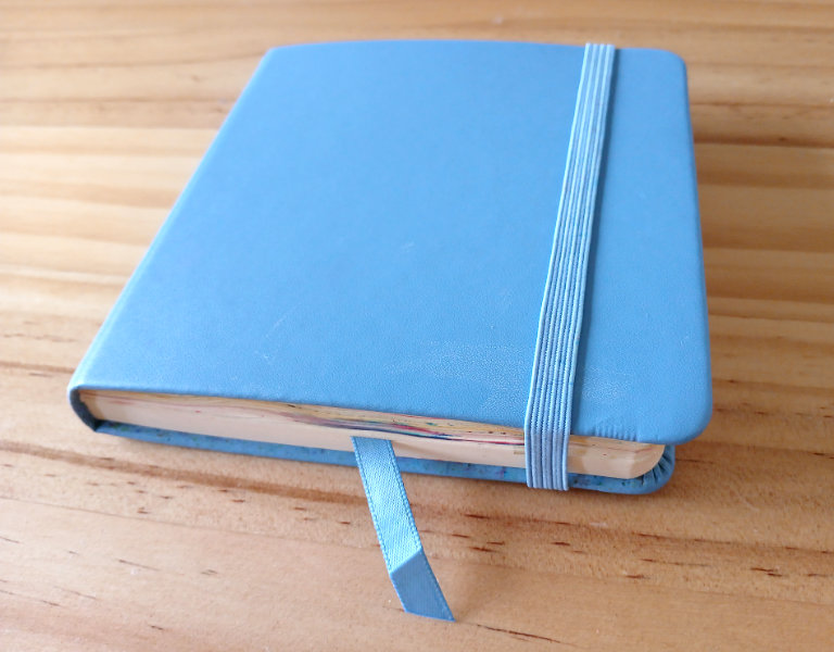 A photograph of Ruth Burton's mixed media sketchbook. It is a small, blue, square sketchbook with an elastic tie and ribbon bookmark.